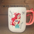 A white ceramic mug featuring a printed illustration of Disney's Ariel the Little Mermaid, with a pink handle and a pink ribbon tied around it, set against a rustic wooden background.