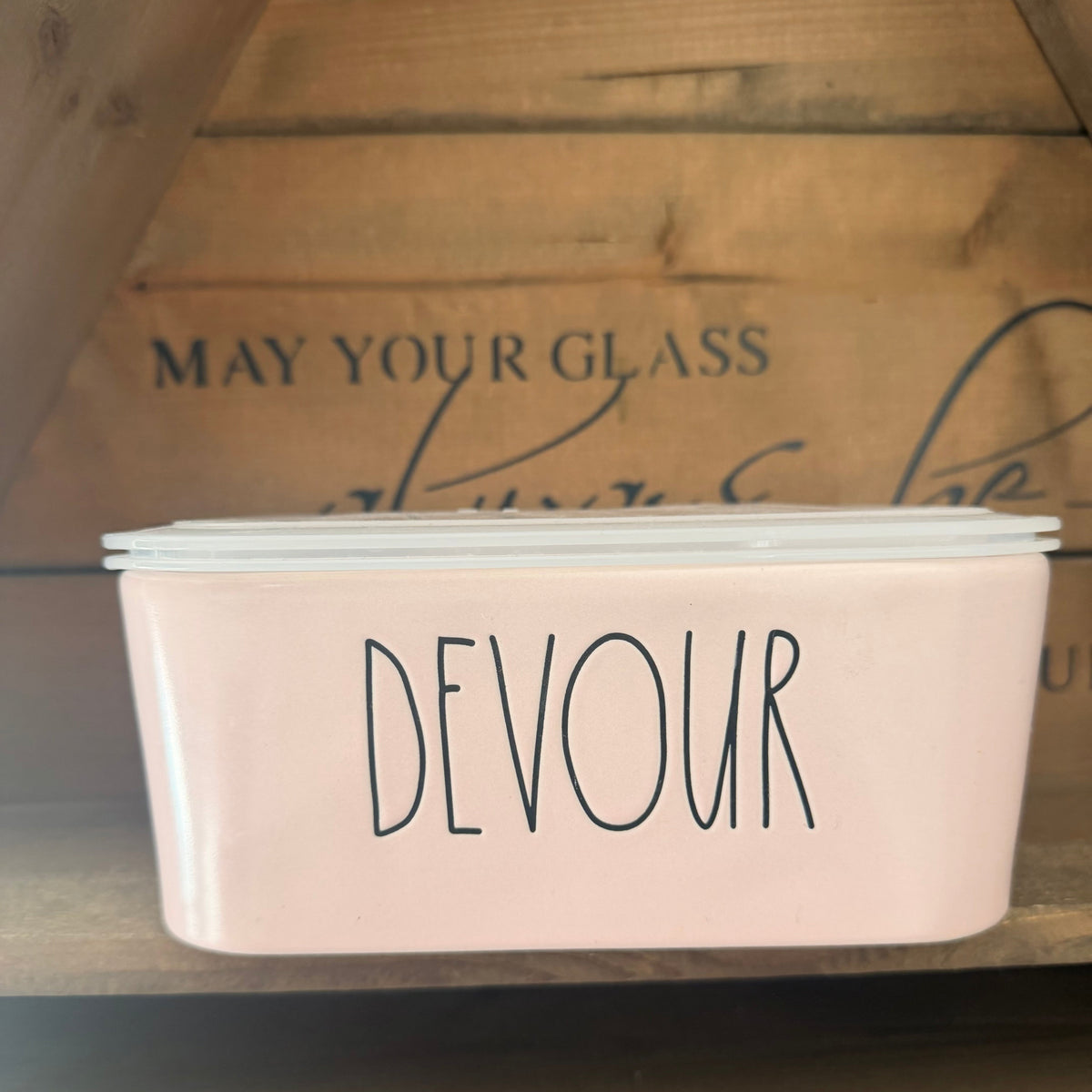 A pale pink ceramic canister with a white lid, featuring the word 'DEVOUR' in black script, displayed on a wooden shelf.